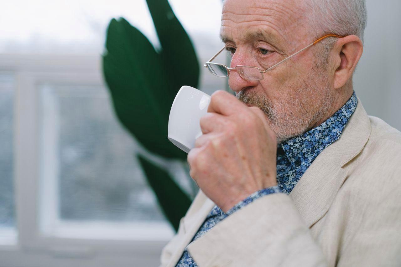 70代の男性がコップ一杯の水をゆっくり飲んでいる、窓辺のリラックスした風景